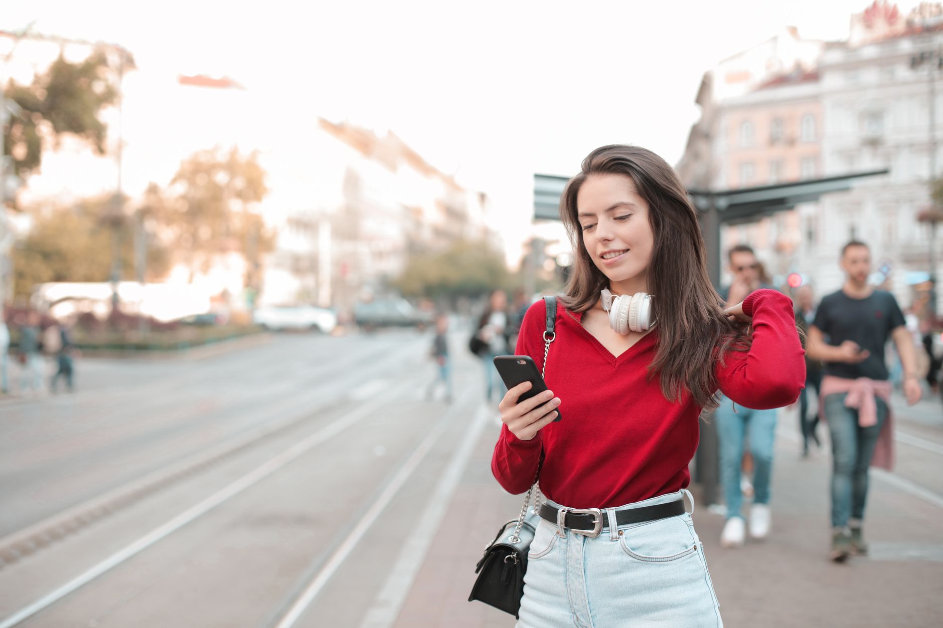 Jeune femme en pull rouge souriant en regardant son téléphone sur un trottoir urbain, des passants flous derrière elle