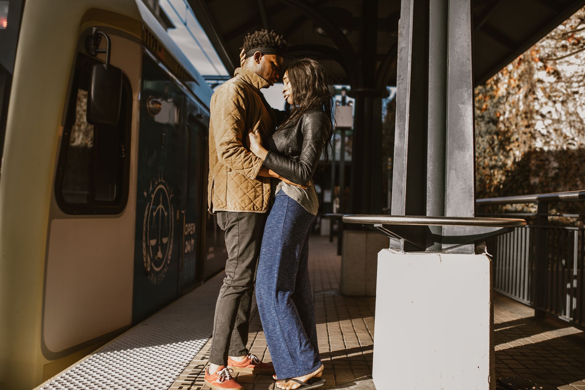 Un couple s’enlace front contre front sur un quai de gare, à côté d’un tramway à l’arrêt, sous une lumière ensoleillée.