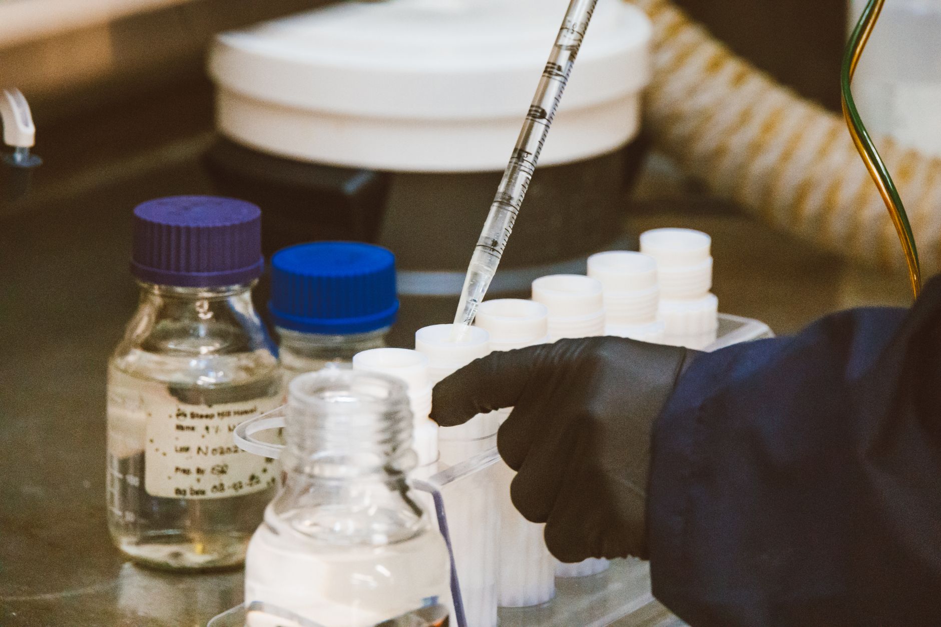 Une personne gantée manipule une pipette graduée au-dessus de flacons et de tubes en plastique dans un laboratoire.