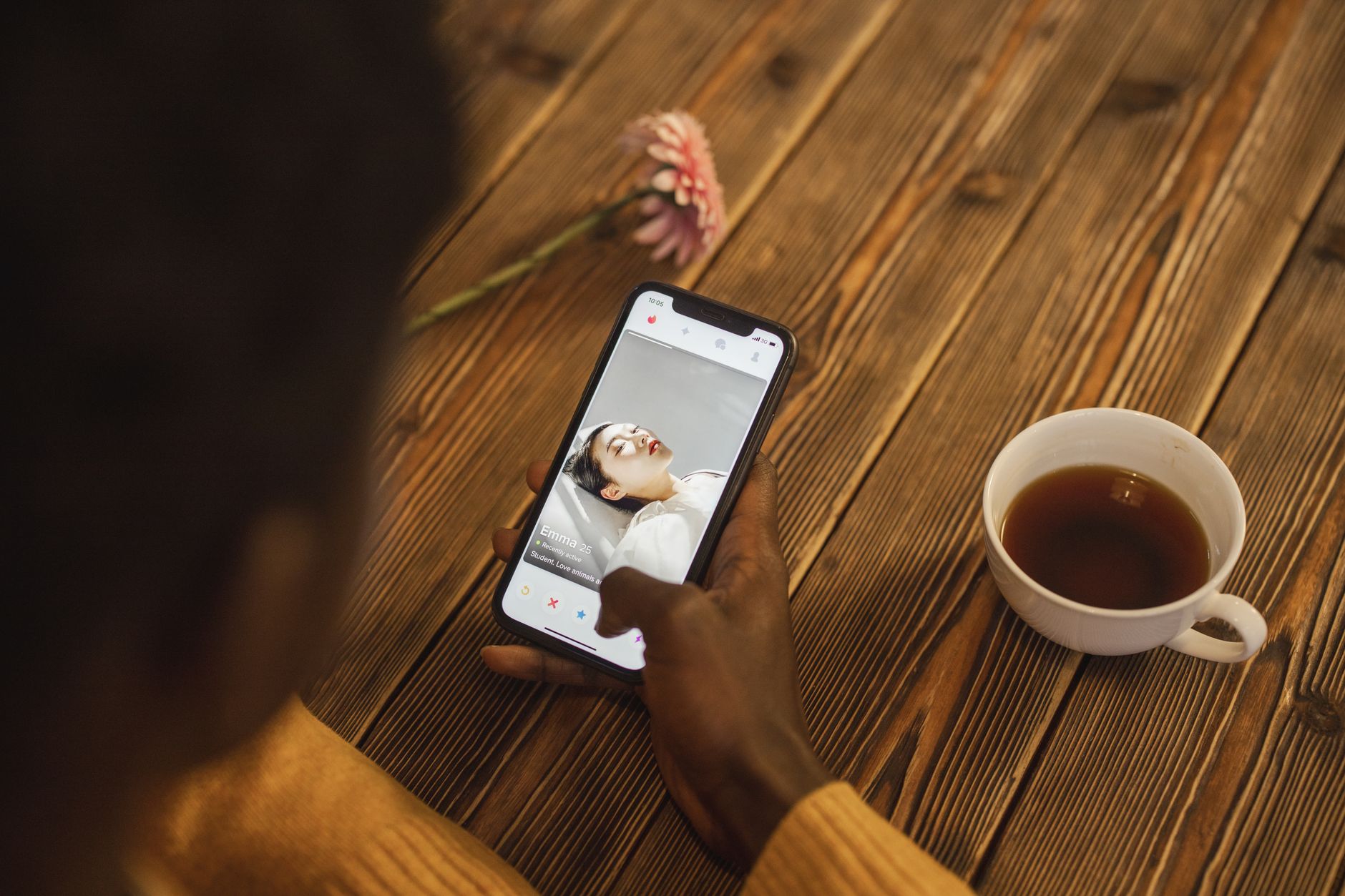 Une personne tient un smartphone affichant un profil sur une application de rencontres, près d’une tasse et d’une fleur rose sur une table en bois.