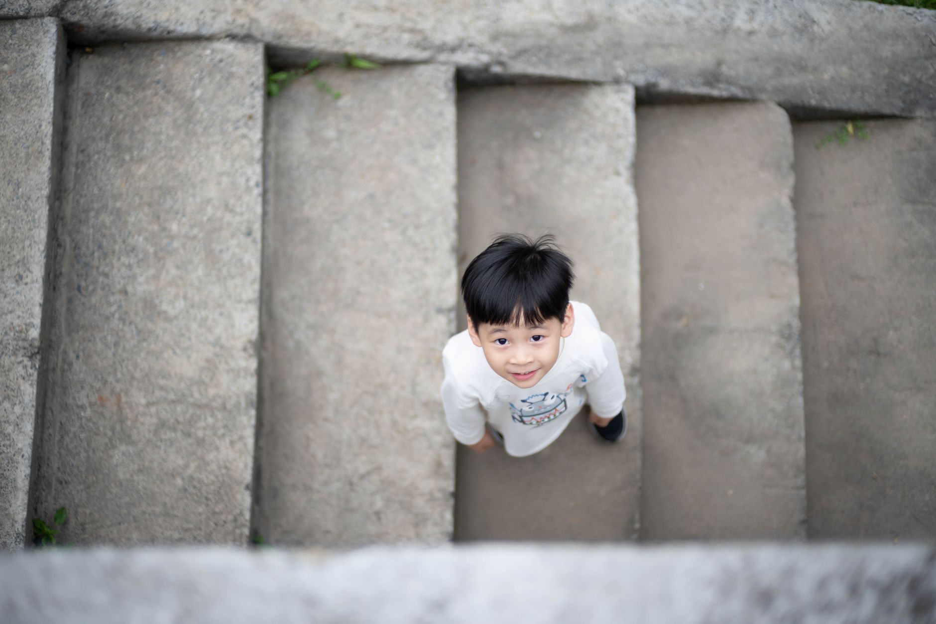 Vue en plongée d’un jeune enfant souriant qui lève les yeux, debout sur de larges marches en béton.